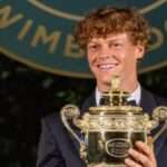 Jannik Sinner vestito elegante posa sorridente con la coppa del Torneo di Wimbledon 2026.